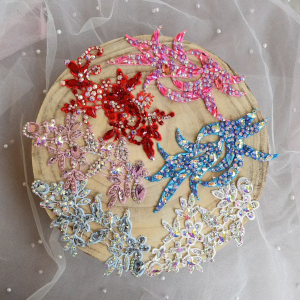 Six embroidered floral and leaf appliqués in red, pink, blue, silver, and white, embellished with rhinestones, arranged on a round wooden slice over a sheer white tulle fabric background. A collection of dance head pieces.