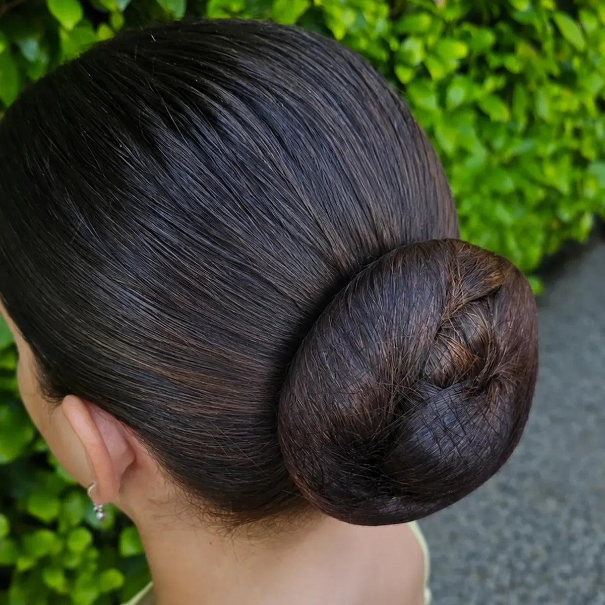 A close-up back view of a woman with her hair in a bun, set against a background of green foliage and a gray path.