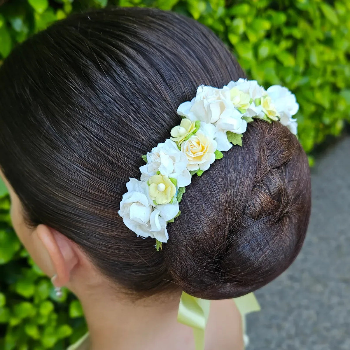 A woman's dark brown hair styled in a low bun, adorned with a white and pale yellow floral hair accessory / ballet bun wrap featuring green leaves, set against a background of green foliage.