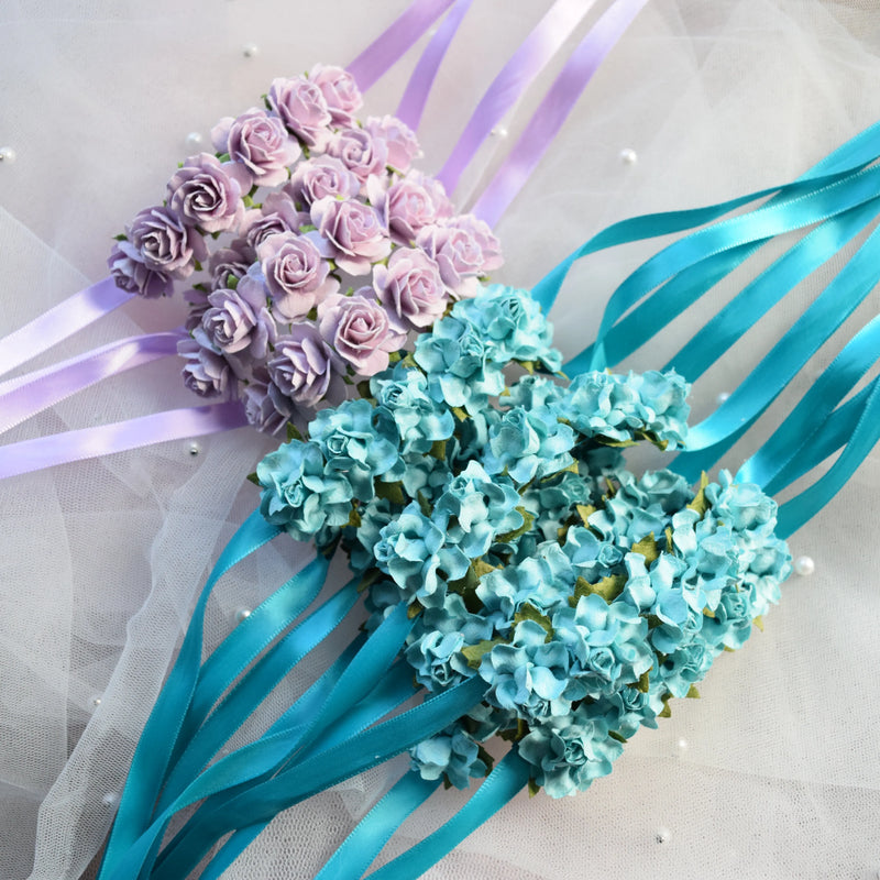 Two styles of ballet bun wraps, one made of light lavender roses with pale purple ribbons and the other made of light blue hydrangeas with turquoise ribbons, laid on a white tulle fabric.