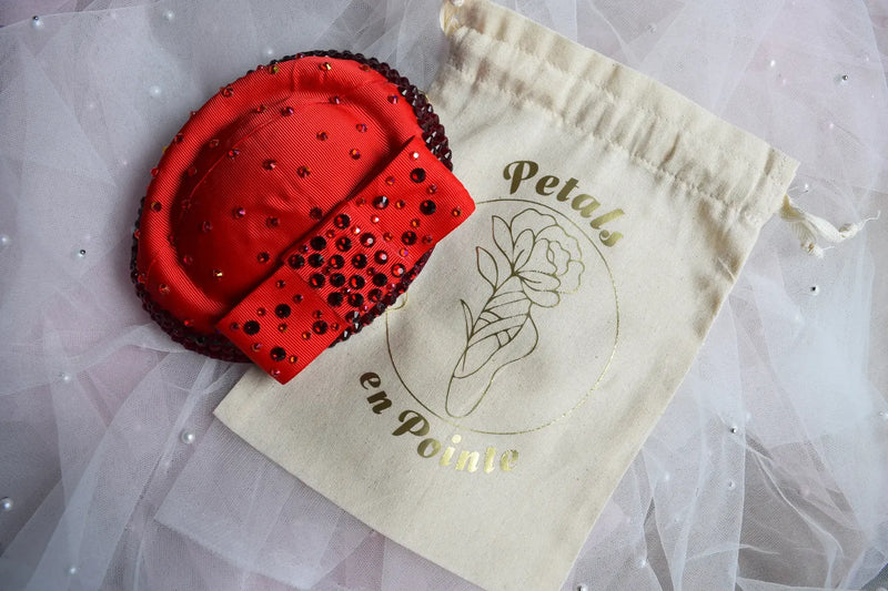 Red custom AB crystal embellished head piece on a beige drawstring bag with 'Petals en Pointe' branding.