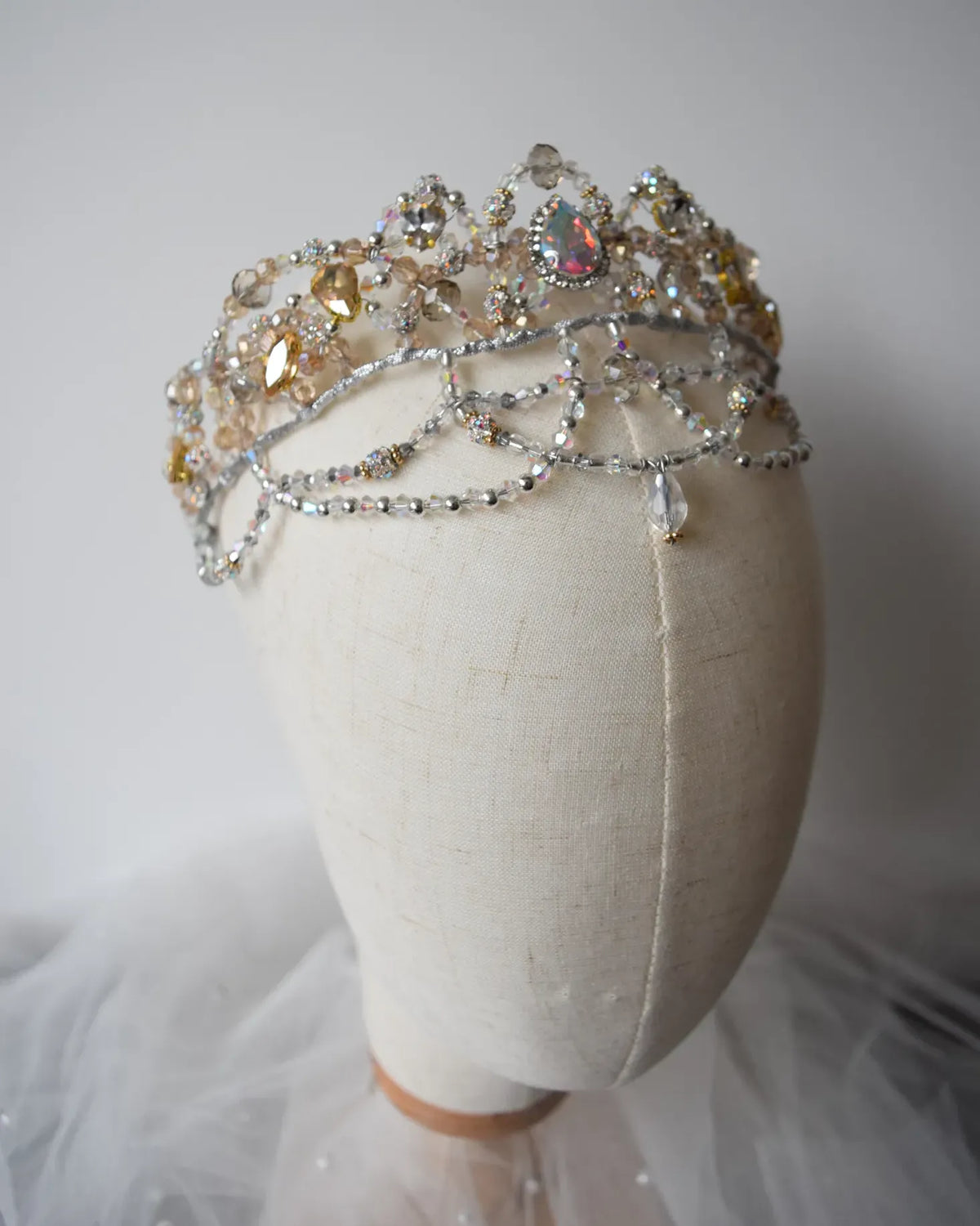 A silver-toned jeweled tiara with clear and iridescent rhinestones, displayed on a cream fabric mannequin head against a soft light gray background.