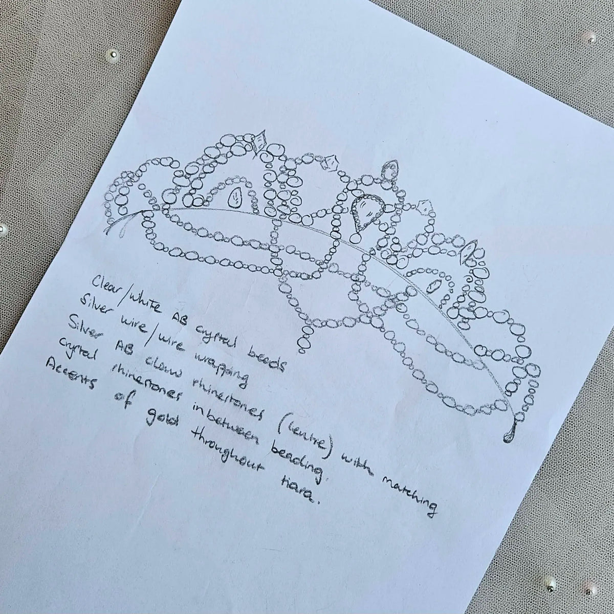 A white sheet of paper with a hand-drawn sketch of a ballet tiara and handwritten notes, placed on a textured beige fabric background.