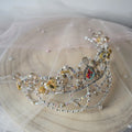 A silver beaded tiara crown adorned with clear crystals, iridescent beads, and gold-toned accents, displayed on a beige fabric surface with a sheer white veil.