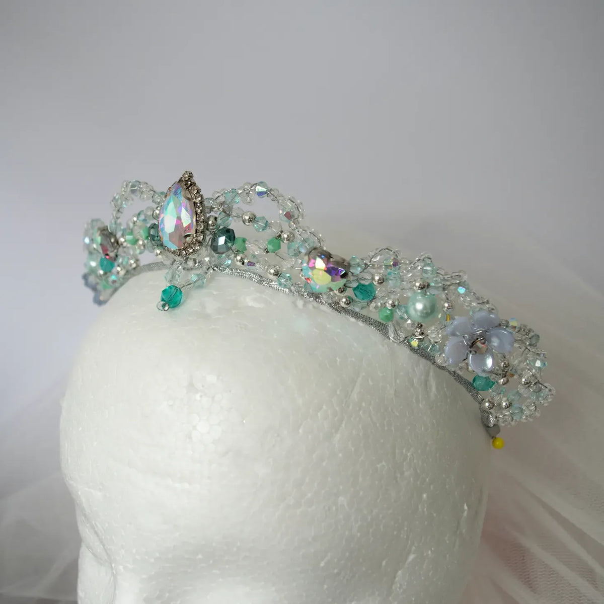 A silver beaded tiara adorned with iridescent, clear, and teal rhinestones, displayed on a white mannequin head against a soft light gray background.
