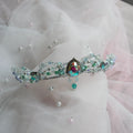 A delicate silver tiara adorned with iridescent rhinestones, teal beads, and a central teardrop-shaped multicolor crystal, displayed on a soft white and pink tulle fabric backdrop.