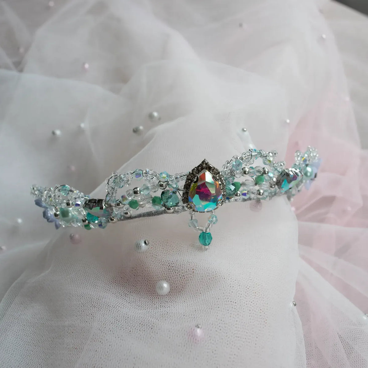 A delicate silver tiara adorned with iridescent rhinestones, teal beads, and a central teardrop-shaped multicolor crystal, displayed on a soft white and pink tulle fabric backdrop.