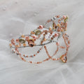 A delicate beaded tiara with rose gold, peach, and silver beads, adorned with faceted gemstones, resting on a soft white tulle fabric background.