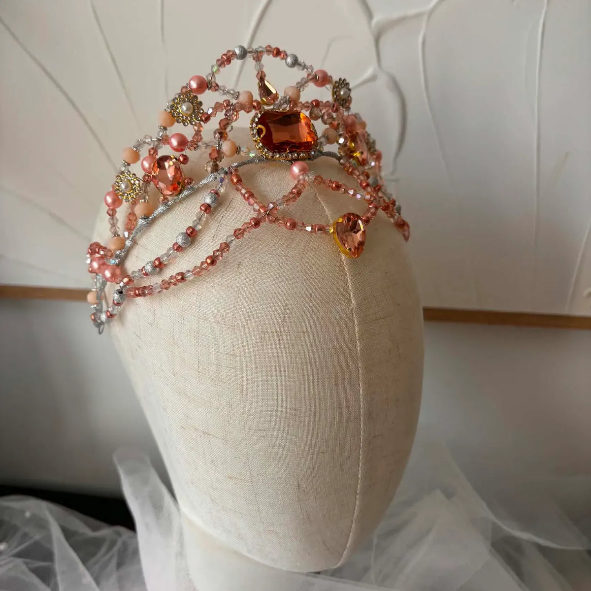 A beaded and jeweled bridal tiara displayed on a cream fabric mannequin head. The tiara features a silver-toned vine design adorned with peach-pink faux pearls, clear rhinestones, and prominent orange and amber faceted crystals.