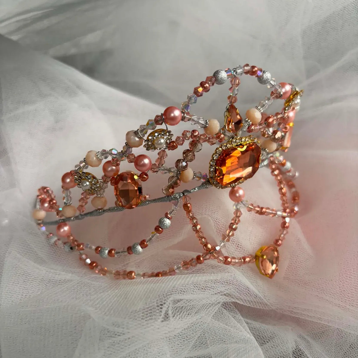 A beaded tiara crown with peach, pink, and amber-colored beads, crystal accents, and a large faceted amber gemstone, displayed on soft white tulle fabric.