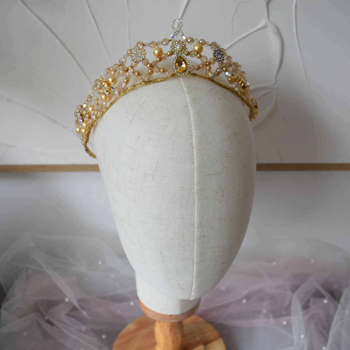 A gold tiara crown embellished with clear rhinestones, white pearls, and a central gold teardrop gem, displayed on a cream fabric mannequin head.