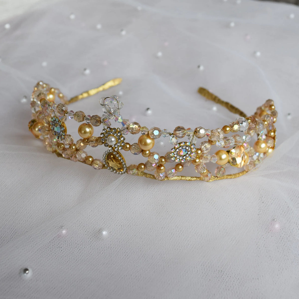 A gold beaded tiara with pearl-like beads, iridescent rhinestones, and a small central crystal accent, displayed on white tulle fabric.
