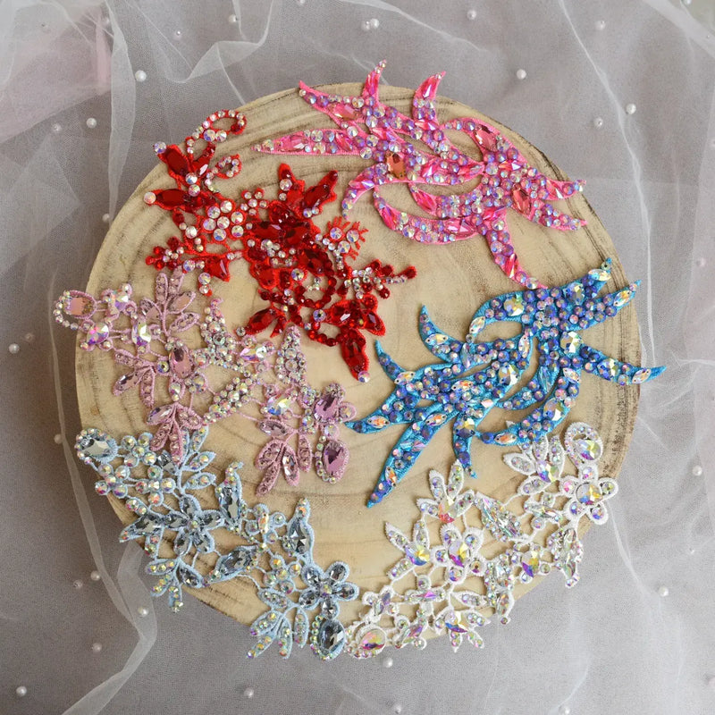 Six embroidered floral and leaf appliqués in red, pink, blue, silver, and white, embellished with rhinestones, arranged on a round wooden slice over a sheer white tulle fabric background. A collection of dance head pieces.