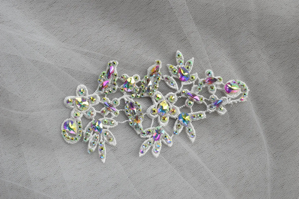 White embroidered lace floral applique embellished with iridescent rhinestones, displayed on a sheer white tulle fabric background.