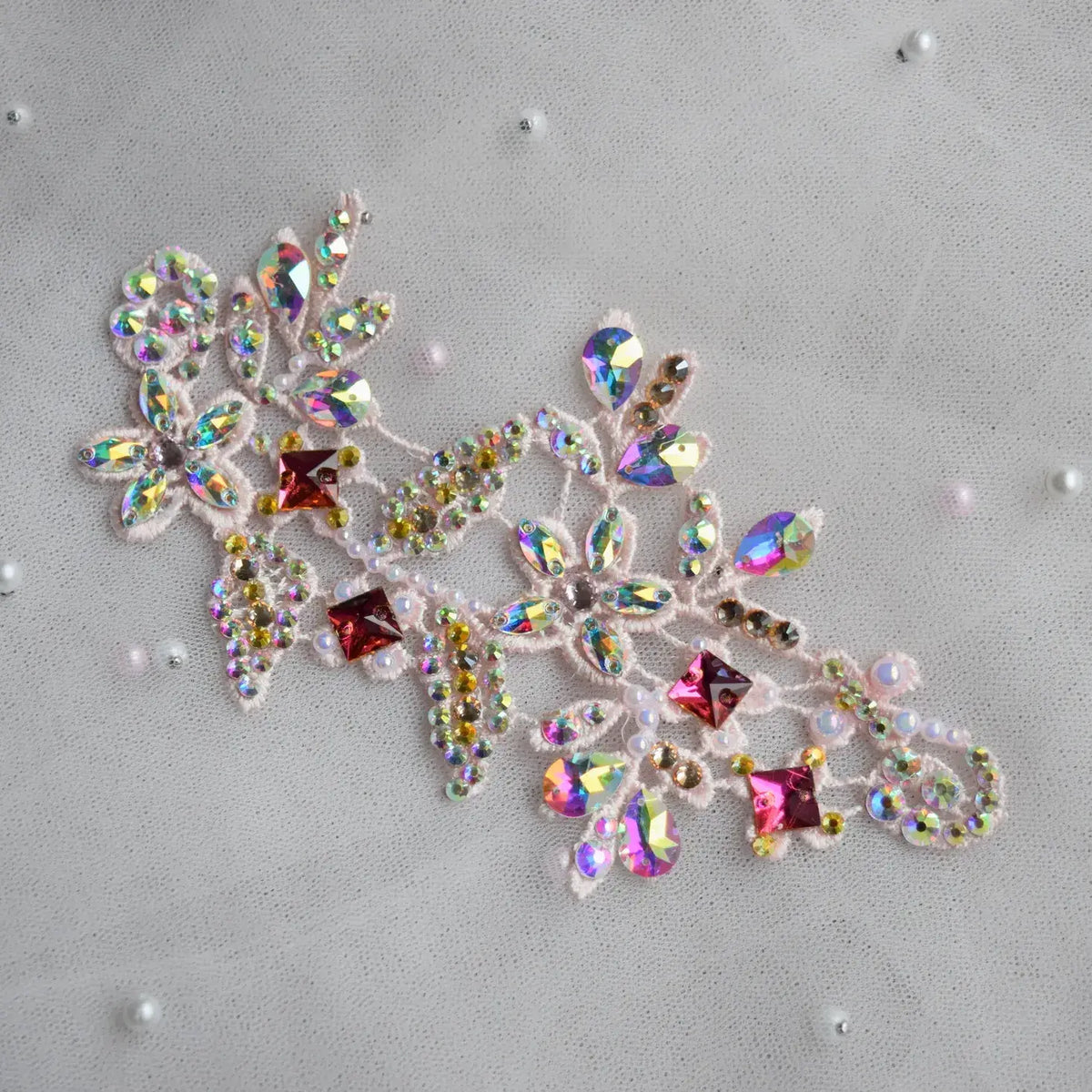 A decorative embroidered lace applique featuring iridescent rhinestones and pink faceted crystals on a light gray fabric background with scattered pearl-like beads.