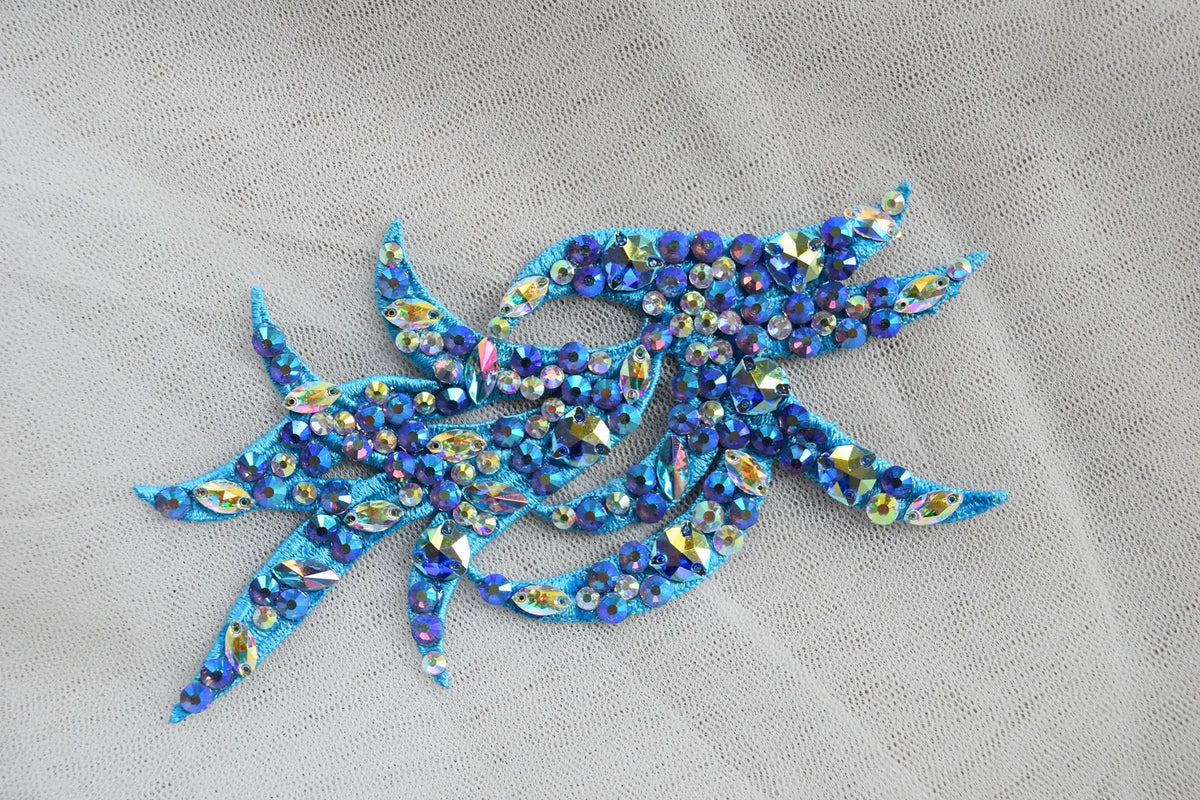 A blue embroidered applique embellished with iridescent rhinestones, placed on a light gray textured fabric background.