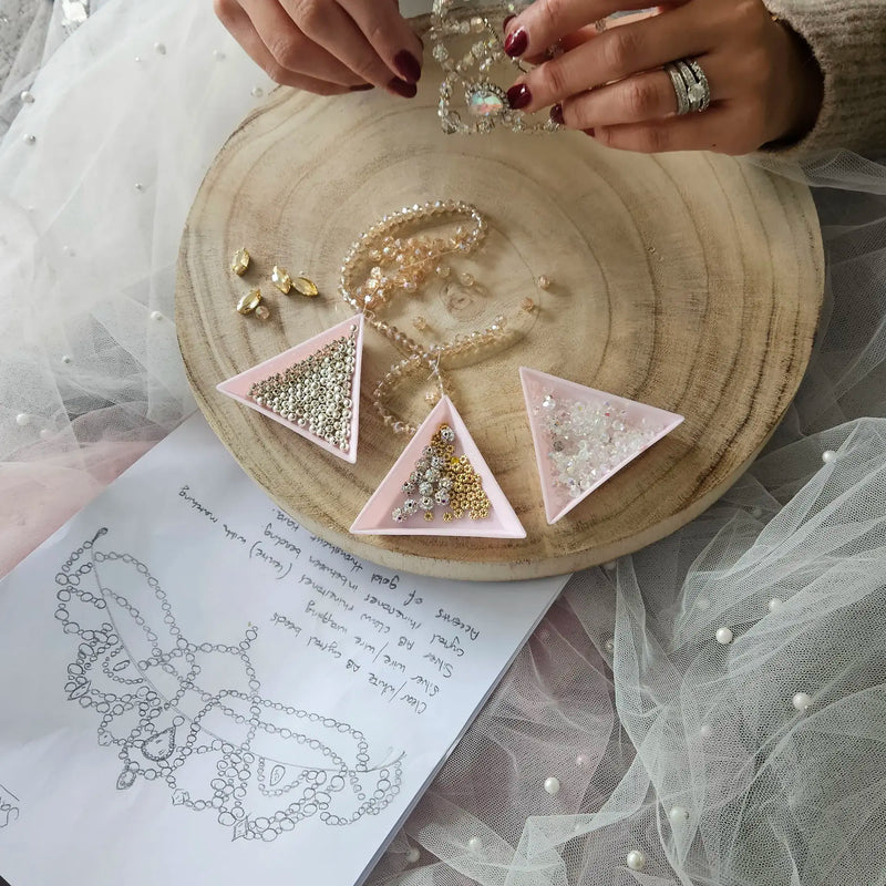 A close-up of hands crafting a custom tiara on a round wooden tray, with pink triangular trays holding gold and silver beads, a pearl necklace, and a sketchbook showing a tiara design below.